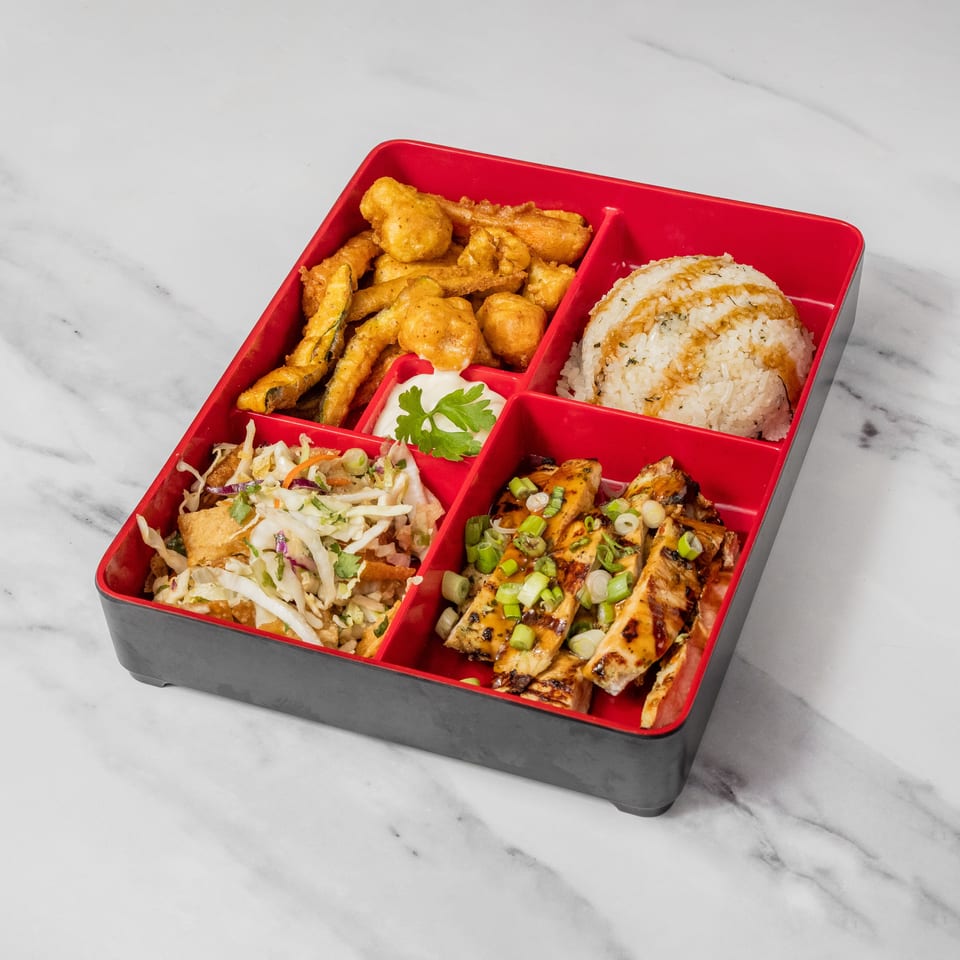 A bento box with fried zucchini, rice, coleslaw, and grilled chicken on a marble surface.