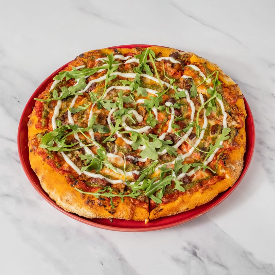 A whole pizza with pesto, tomato sauce, arugula, and a white sauce drizzle on a red plate.