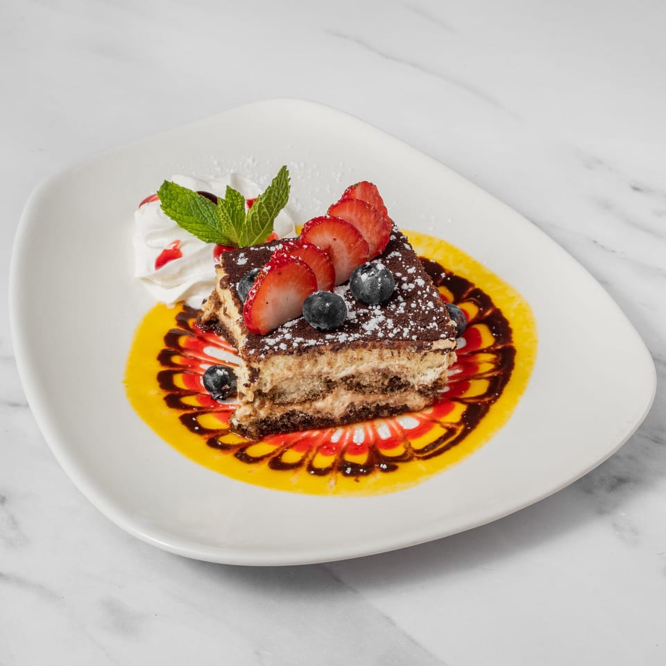 Tiramisu slice with strawberries, blueberries, whipped cream, and decorative sauce on a white plate.